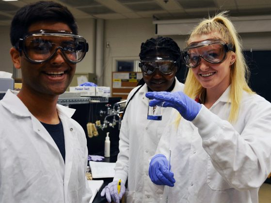 3 students in lab gear holding up a beaker