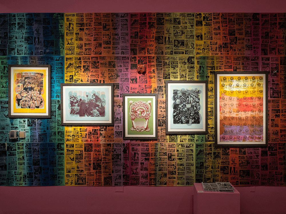 A wall from an art exhibition shows five framed prints set against a repeating pattern of black and white images set against rainbow colors. The artwork is from the exhibition With Care by artist Nicole Marroquin and includes black and white photographs mixed with bright colors.