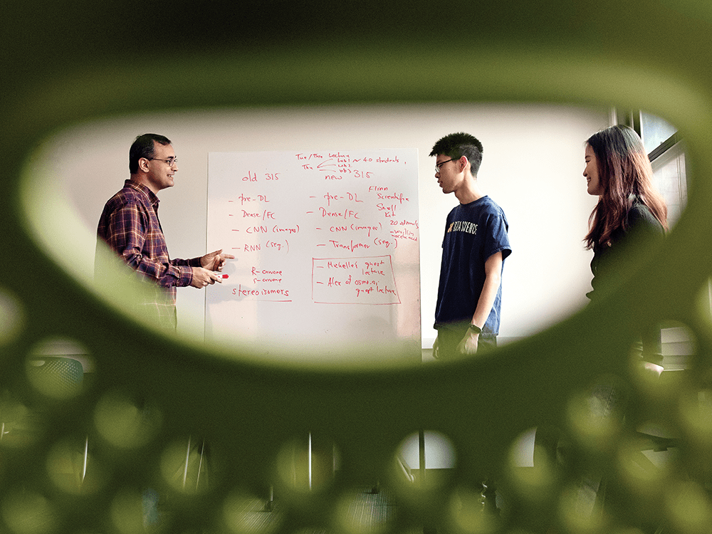 In a photo taken through a large, oval hole in a green chair, Professor Ambuj Tewari stands on the left of a whiteboard with red writing on it. He points at the whiteboard as he smiles at students Yikun Han and Rui Nie, who are on the right side of the whiteboard.