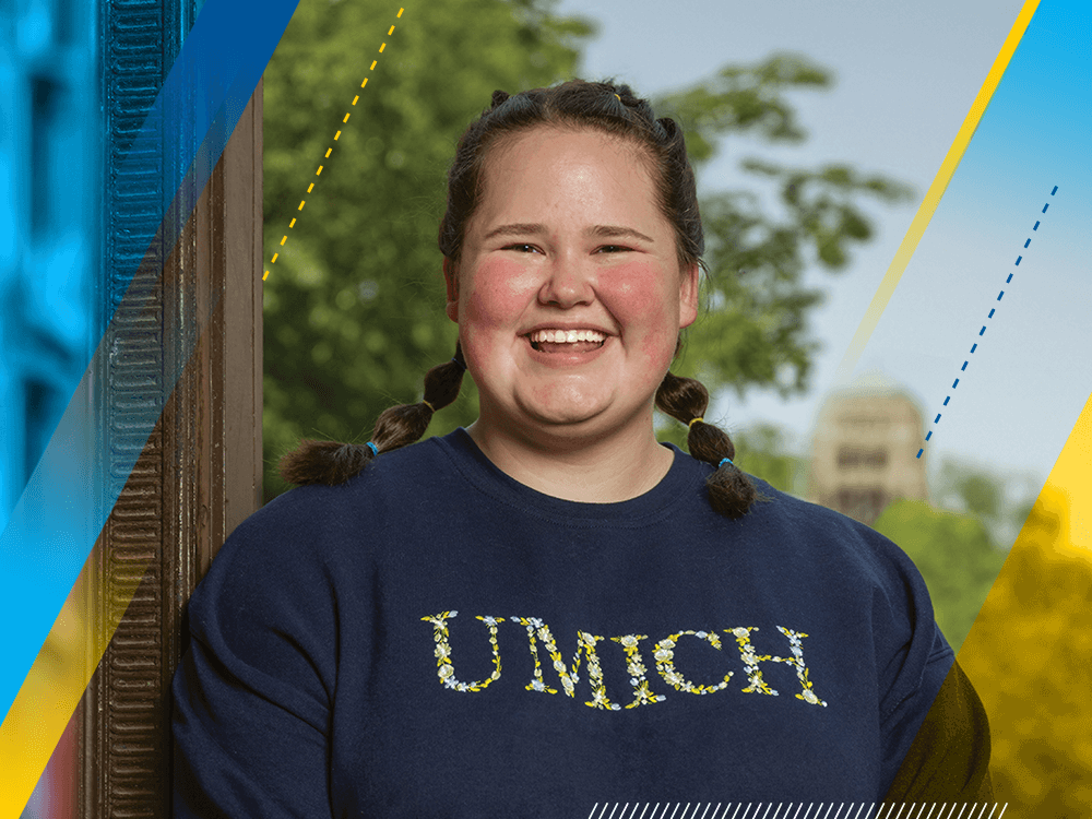 A female college student with pigtails smiles broadly. She is standing outside. She wears a blue sweatshirt with “UMICH” on it.