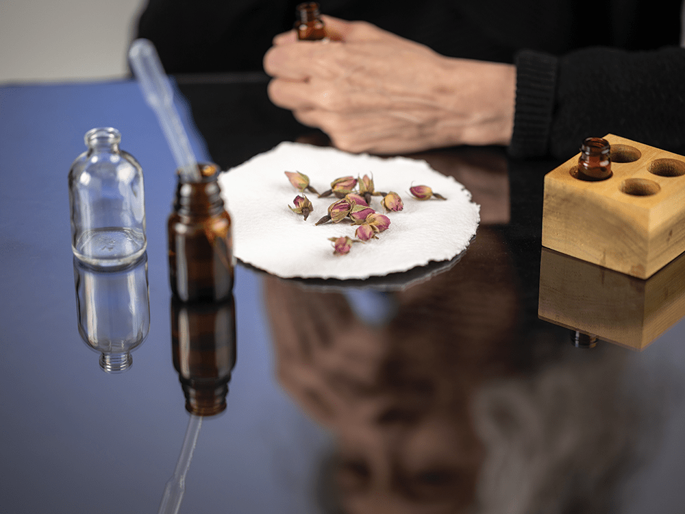 Looking down from above a clear table, Michelle Kydd holds a small, brown scent bottle and is out of focus in the background. On the table, a circular piece of paper with rose buds on it is in the center, in focus. The photo also includes two more scent bottles, a pipette, and a portion of a wooden container.