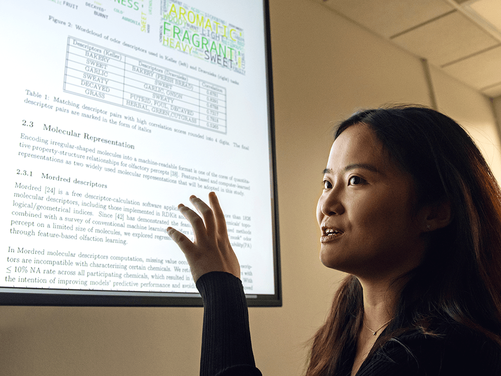 Student Rui Nie stands next to a projection of a scientific paper, with one of her hands raised and the light from the projection shining on her face.