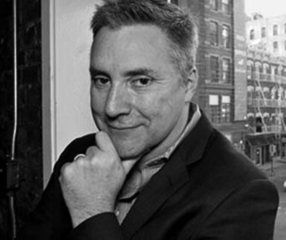 Black and white headshot of Joel Tractenberg, Honors Alumni Council Member, from the shoulders up. He is posing with his hand on his chin wearing a black suit. He is posed in front of a window with a city landscape in the background. 