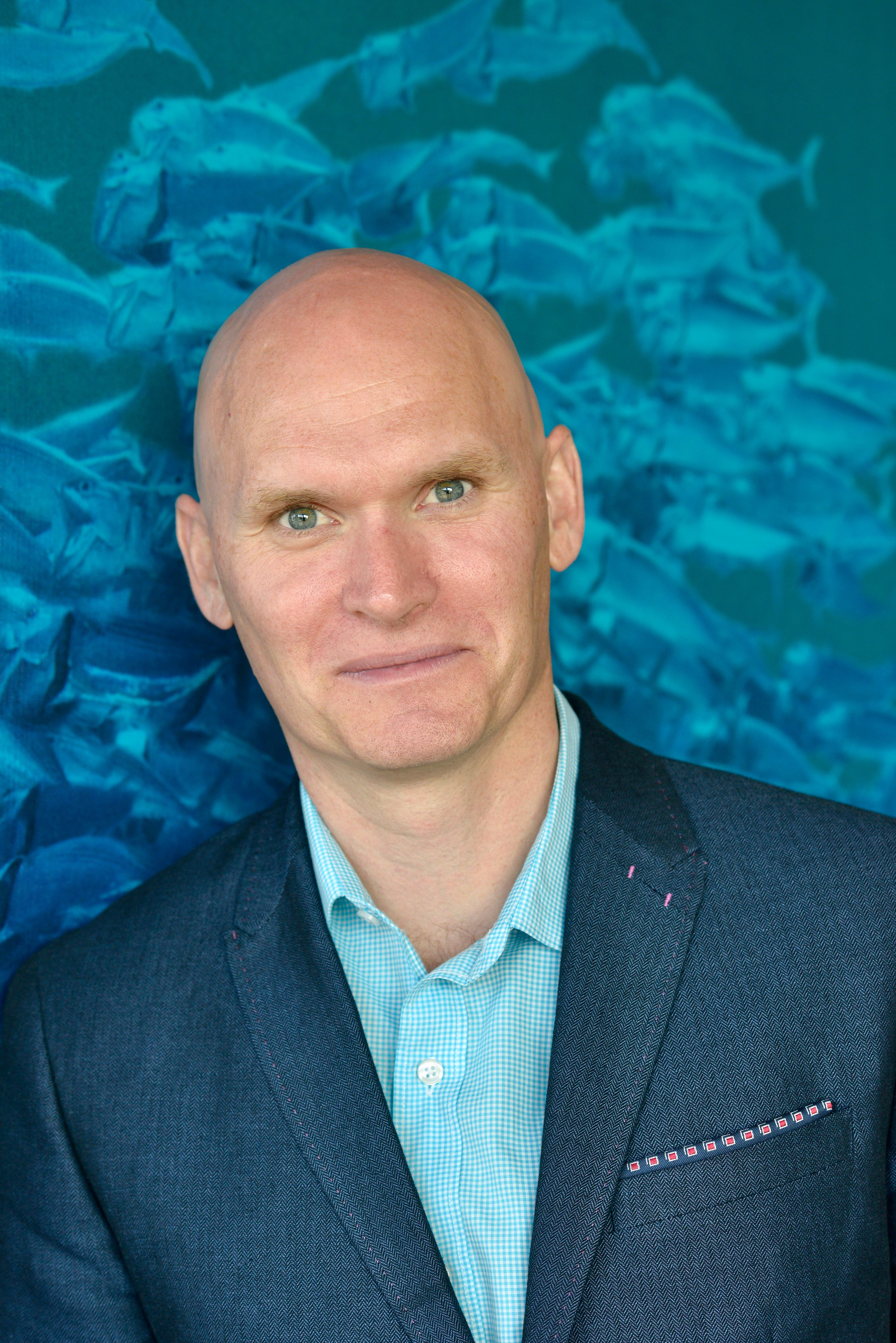 Image of Anthony Doerr from the chest up wearing a blue suit with a turquoise background. 