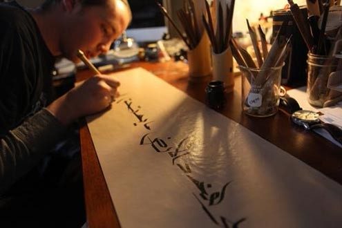 A person leans over a desk writing Islamic calligraphy in dark ink on white paper.