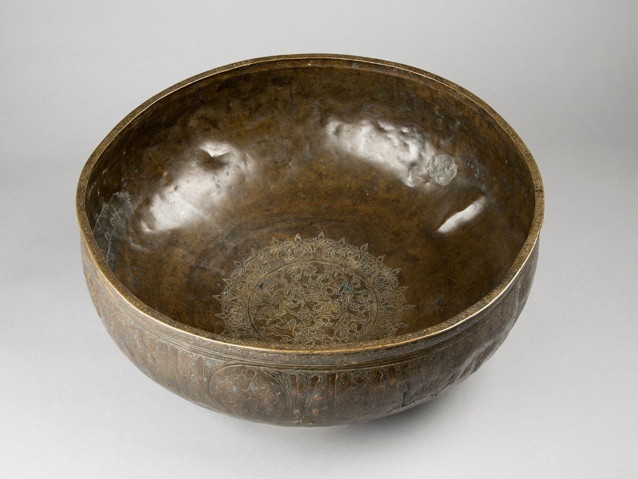 Brass bowl with a fish and pond pattern carved at the bottom of the object’s interior.  Additional inscriptions are visible around the bowl’s exterior. 