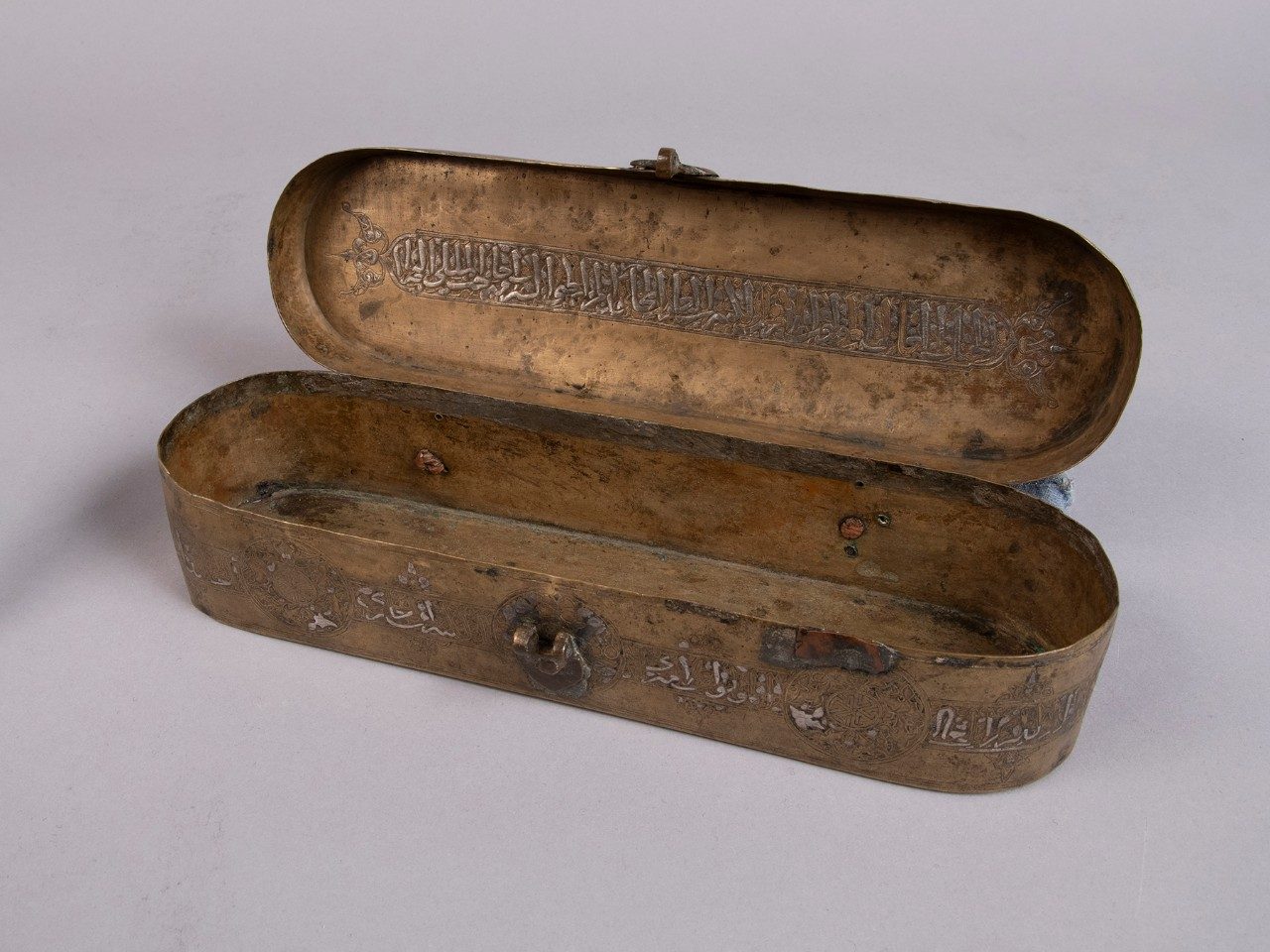 Oblong hinge-top brass box with a clasp on its front. Arabic inscriptions line the exterior of the case, and the open top reveals more writing.