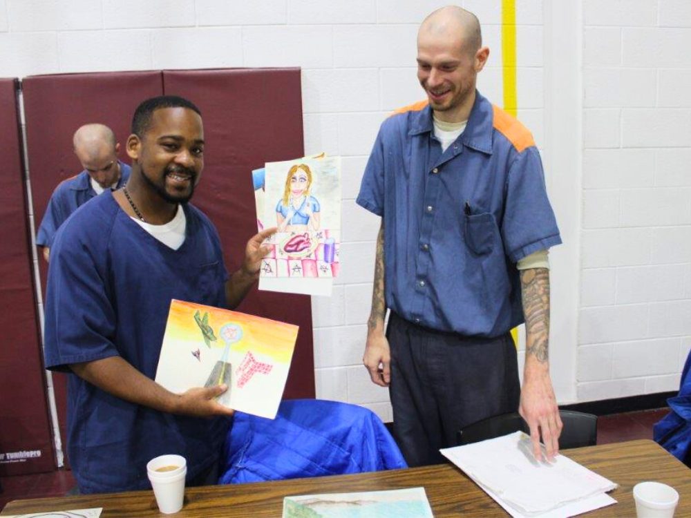 Two incarcerated men. One holding up artwork he created. The other smiling next to him 