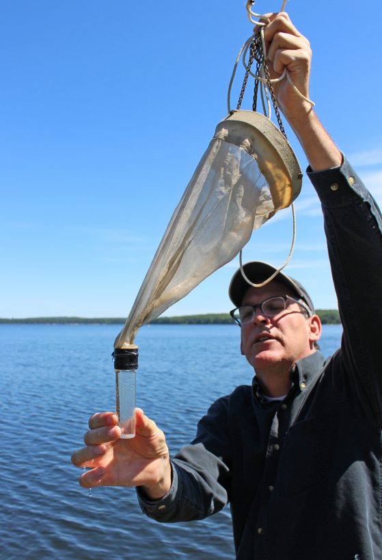 A man holds up a plankton tow, which comprises a cone-shaped net with a small glass bottle on the end.