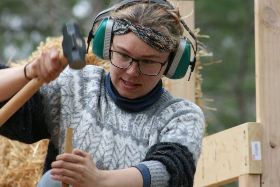 A female student, wearing a headset, hammers a dowel during construction.  
