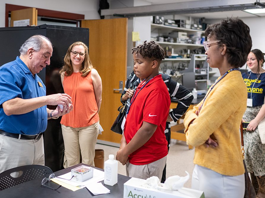 A man in a blue shirt shows a woman in orange, a boy in red, and a woman in yellow research being conducted in one of Homer A. Neal's new labs.