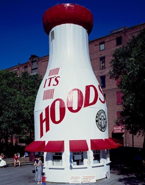 a white kiosk with red awnings over several windows, the kiosk is shaped like a large milk bottle with a red cap and red text that reads "Hoods"