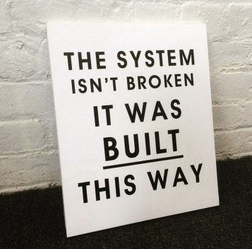 Black text on white board leaning against a white brick wall.  Text reads: "The system isn't broken it was always built this way"