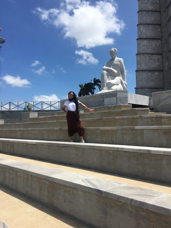 A female student standing on a large set of stone stairs in front of a white statue of a woman kneeling. The sky behind her is blue with a few clouds, but very sunny. 