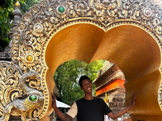 Photo of Harrison, a 6 foot black male student, smiling casually for photo under an ornate gilded and bejeweled gate entryway in Thailand. 