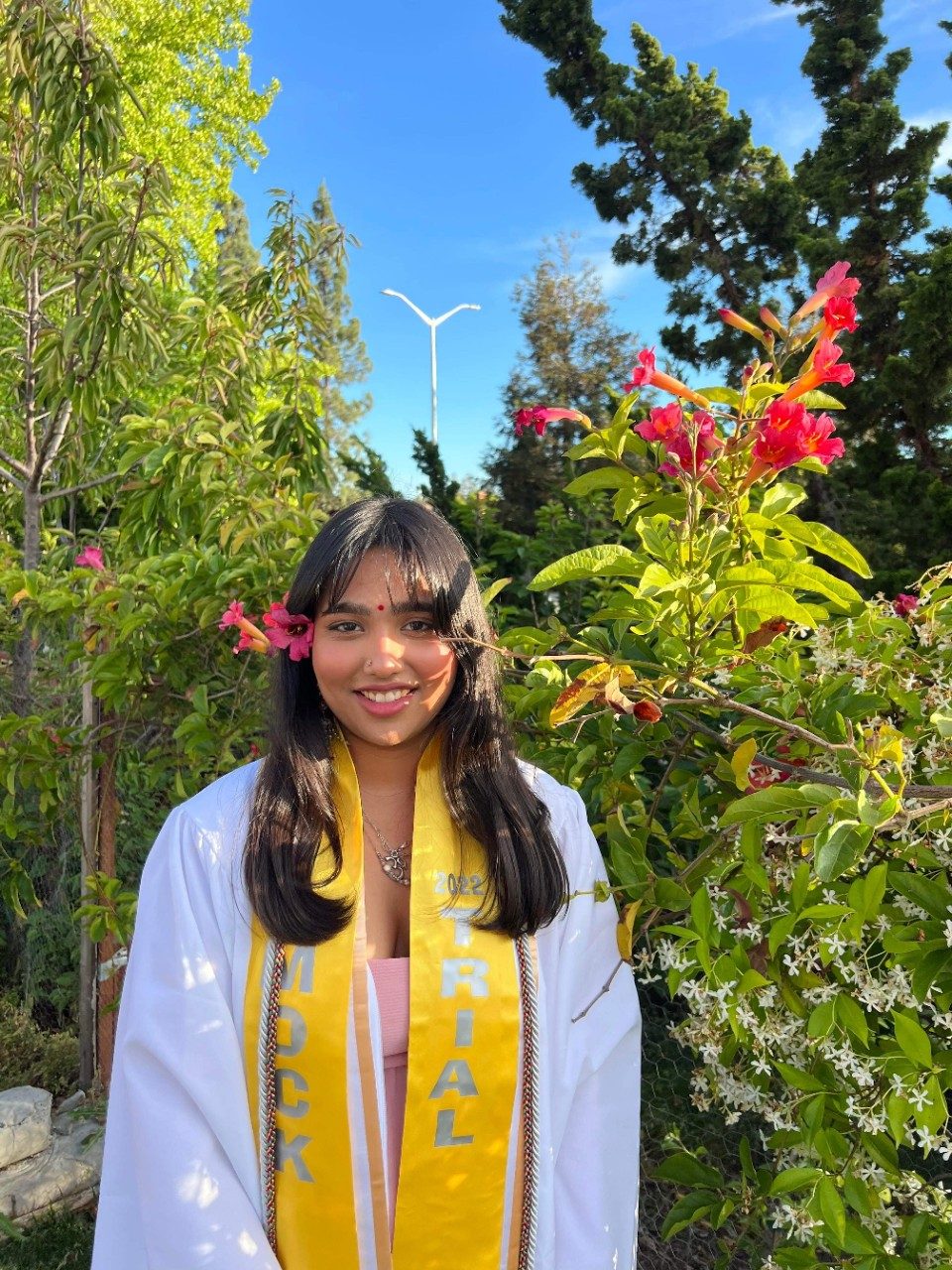 Photo of Devanshi in a white graduation gown with yellow sash against beautiful trees and flowers. Link to submission.