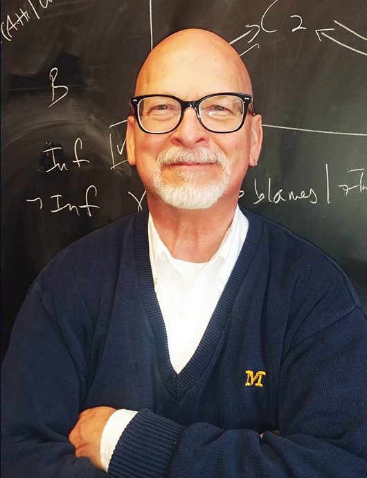 Charlie Doering with arms crossed over chest wearing Michigan M sweater over white collared shirt - with slightly mischievous smile against blackboard with math on it