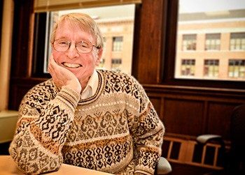 John Holland smiling at table with chin in right hand. Patterned brown sweater with collared shirt underneath.
