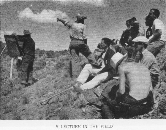 Black and white photograph of surveying class in hillside near Camp Davis, central figure standing and gesturing to the distance, seated group of students in foreground.