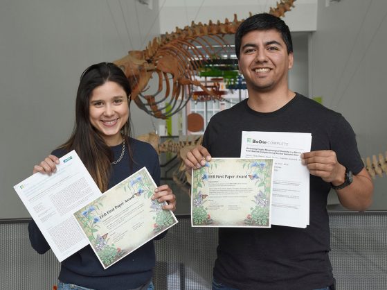 Sara Colom and Peter Cerda hold up their certificates and copies of their papers in the musem atrium with prehistoric whales in the background