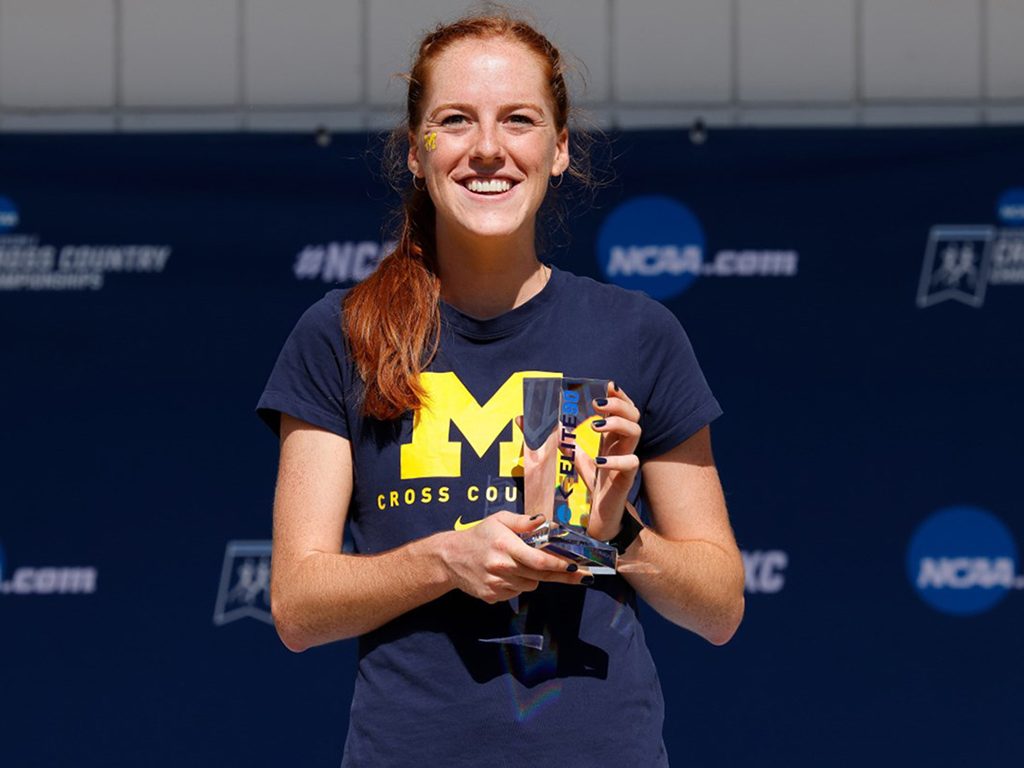 Alice Hill holding her Elite 90 trophy in front of an NCAA banner and wearing her University of Michigan cross country T-shirt