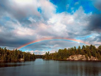 Rainbow over the Michigamme Highlands, full rainbow arcing over a lake, rocks and pine trees on either side of the shore, blue sky with clouds