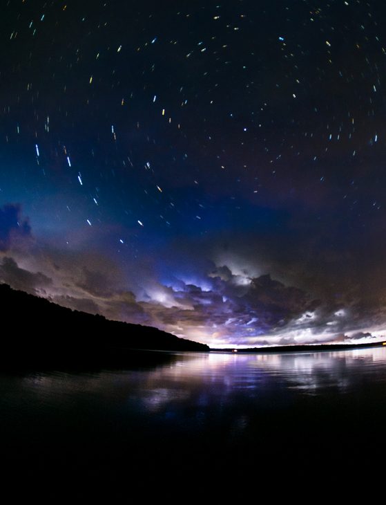 Fifteen minutes at the Biostation, U-M Biological Station. Image credit: Will Weaver. Starry night sky over a lake and hilly shore on one side, stars have short trails through extended exposure photography, lightning lighting the horizon