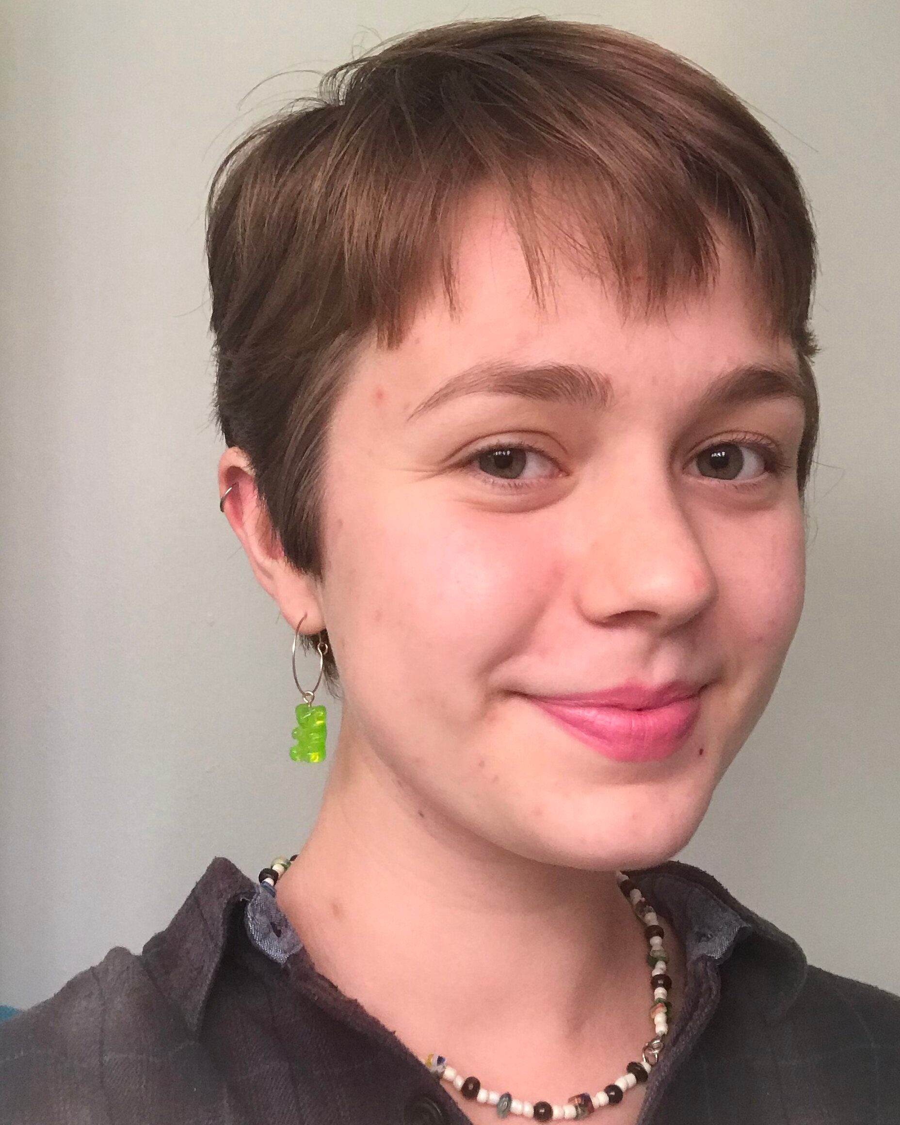 Person with short straight light brown hair smiling wearing a brown shirt and a beaded neacklace and a green gummy bear earing