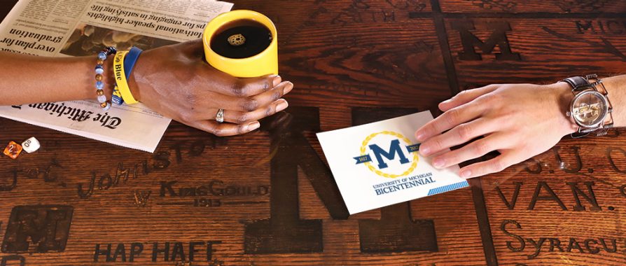 Two hands on a table that has block M, names, and other graffiti carved into its surface. One hand it touching a 20 questions with LSA playing card. The other hand is holding a cup of coffee in a yellow mug. 
