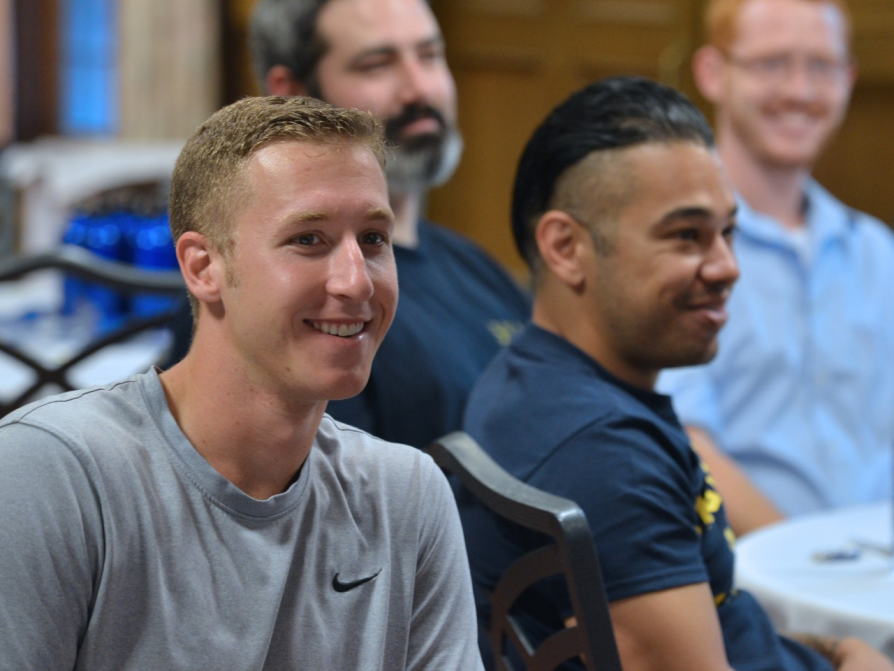 A classroom photograph in which two men are primarily featured. They are both smiling and attentive. Other men are included in the photo in the background.