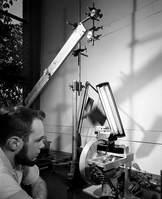 A black-and-white photograph of a man leaning intently toward a mechanical-looking device with his left elbow on the table.