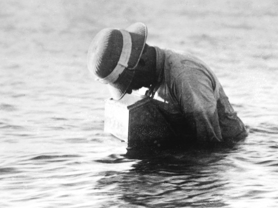 A black-and-white photograph of Reighard standing in waist-deep water taking photographs within the watertight box he had designed.