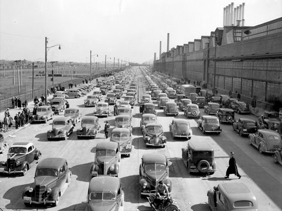 A black-and-white photograph of several very long lines of cars outside the River Rouge plant. The street is adjacent to the plant, which towers above it. Seven slender smokestacks reach up from its roof to the sky. The photo is taken from above, and you can see a few people walking beside and between cars.