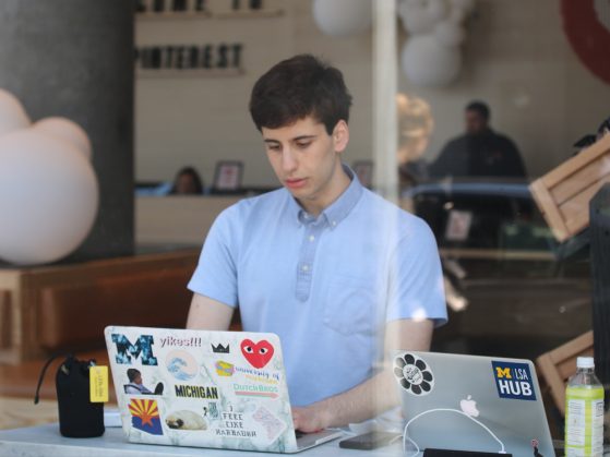 A photograph taken through a window of a young man wearing a blue button-down shirt working at a laptop in a public space, such as a cafe. The laptop is covered in stickers.