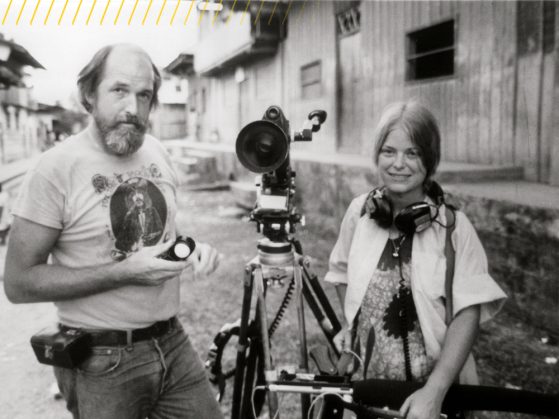 A black and white photo of Maureen Gosling wearing a large pair of headphones. She stands next to a film camera and Les Blank.. 