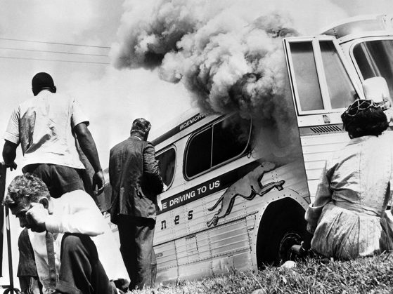 A black and white photo of a Greyhound bus with a plume of smoke billowing out its door. A woman sits in the grass looking at it. Another man sits in the grass leaning away from the smoke. Two men are standing facing the bus. The Greyhound slogan, Leave the driving to us, is partially legible on the bus's side.