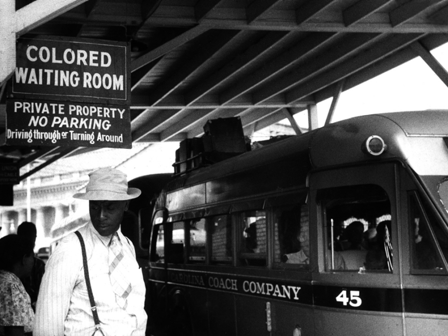 In a black-and-white photograph, a black man stands beneath a sign that says Colored Waiting Room in large letters. Below, in smaller letters, the sign says private property. No parking driving through or turning around. The man is wearing a white hat, a striped shirt, a striped tie, and suspenders. He is looking down to his right. Behind him, seen over his left shoulder, is a bus. 