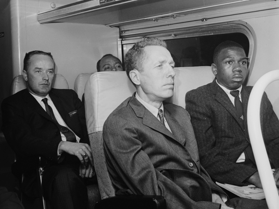 A black and white photograph of four men sitting on a bus. James Peck sits on the aisle and Charles Persons sits next to him. Both men are wearing suits and staring straight ahead.