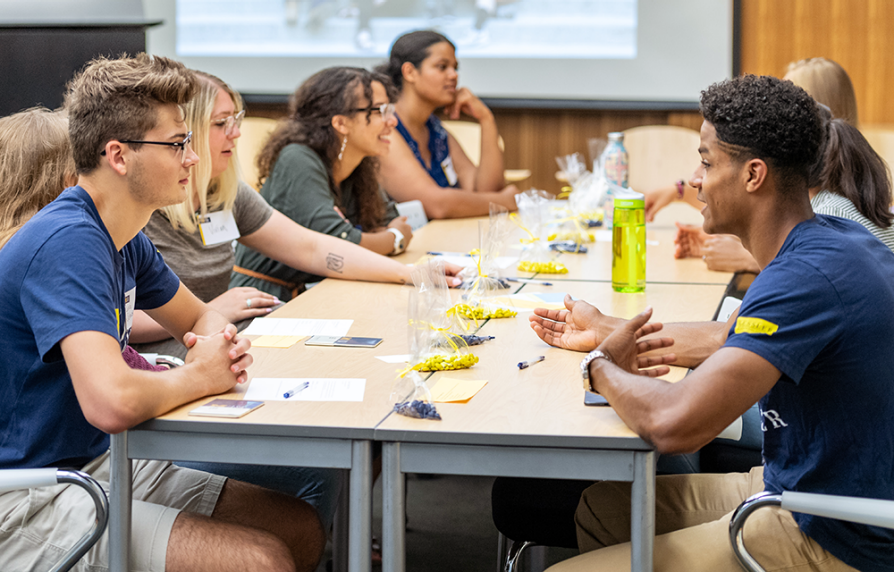 Students sitting side by side at a long table, leaning forward to talk to each other. There are plastic bags of blue and yellow M&Ms in the center of the table, which the students are using as they participate in an interactive game.