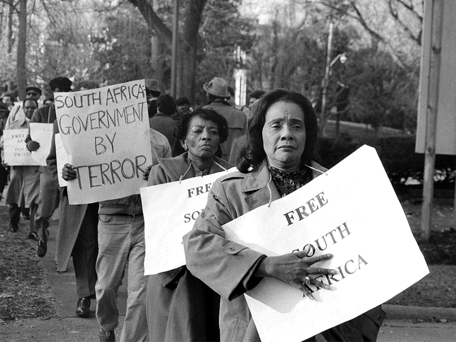 Black and white photograph of a line of marching protesters carrying signs that say free South Africa and South Africa government by terror. The marchers are wearing warm coats and the signs are hung by strings around their necks as if they were necklaces. Coretta Scott King is at the front of the line. She's looking into the distance and is holding the sign against her body with her open right hand.