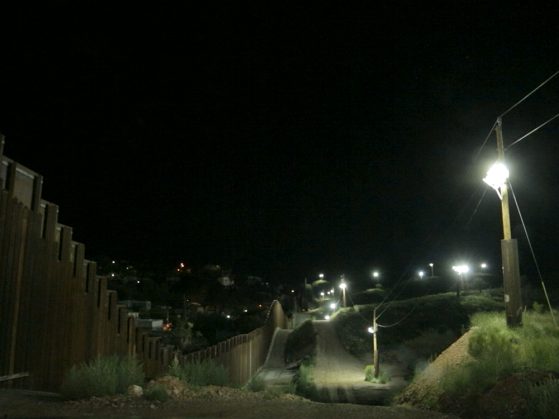 A photograph of the border at night. The dark sky is punctuated by a series of distant streetlights,and fainter lights can be seen of a city below the road. The fence rises and dips along with the road.