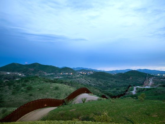 A daytime photograph of the border fence, which runs along a dirt road. There are lush green hills, a bright blue sky, and foothills in the distance.