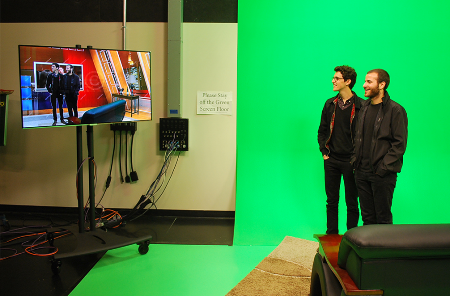 Two young men stand on a green floor in front of a green wall. They are looking at a television screen that mirrors them, but shows them in a room similar to a television news studio.