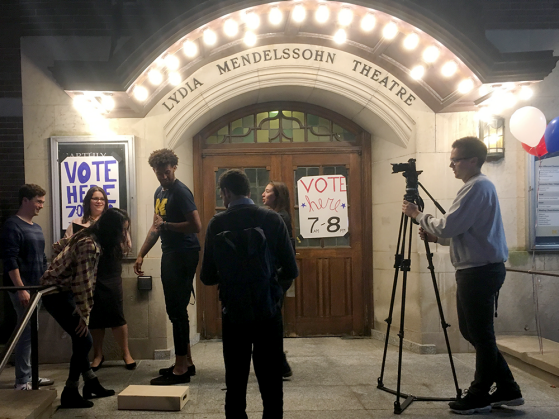 Students filming a PSA outside of Lydia Mendelssohn Theatre. Five students stand outside the building, which has a poster on its door that says VOTE *here* 7am-8pm and another on its announcement board. Another student stands behind a camera filming them. Three helium balloons are tied to the railing: one red, one white, one blue.