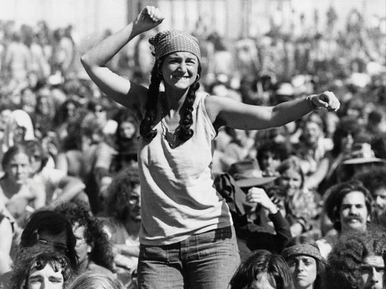 Black and white photo of woman in a crowd on someone's shoulders with her fist in the air.