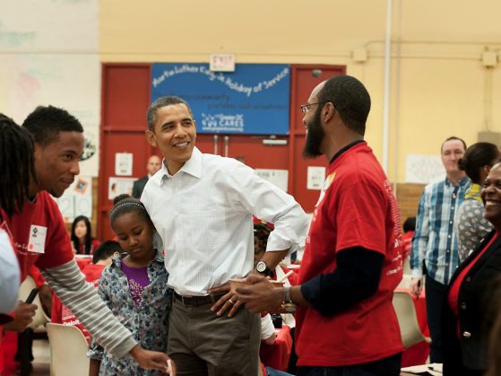 L. Trenton Marsh, wearing a red volunteer shirt, stands in a school cafeteria talking to President Barack Obama. President Obama is looking at Marsh over his left shoulder and also has his right arm around a young girl. 