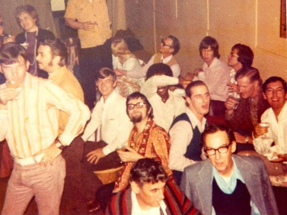 A photograph of about 15 patrons of the Up Stairs lounge. Some are sitting at tables holding cocktails; others are posing by holding a hand on a cocked hip. They are all men.