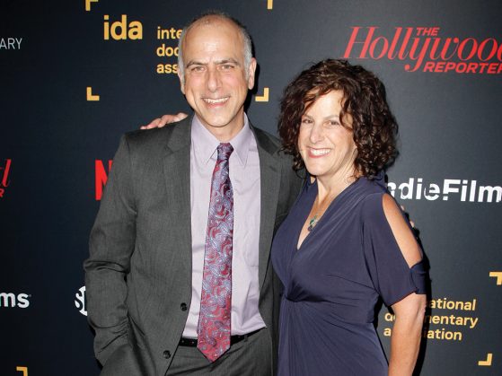 Jody Becker, wearing a blue formal dress, stands with Dan Habib, who's wearing a suit and a tie, in front of a black screen that has the names of different film organizations, such as International Documentary Association, and film publications, such as Hollywood Reporter, written in different colors and fonts across the screen.