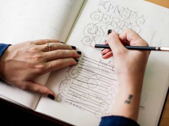 A woman's hands on an open sketch pad. The left hand holds the pages open and the right hand holds a pencil. The page has sketches for ideas for a wine label, such as floral script and curlicues.