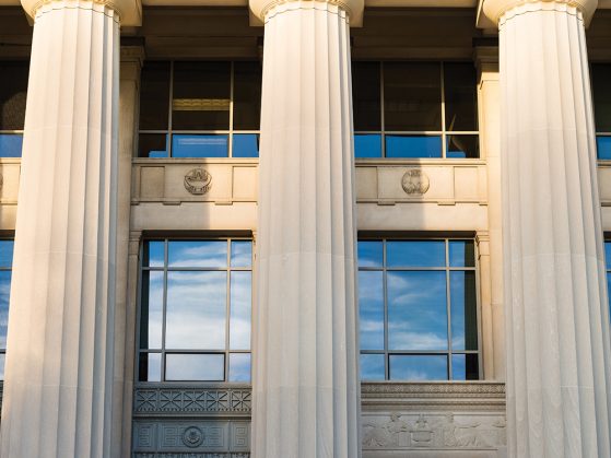 A photograph of the pillars of Angell Hall in which the windows reflect a bright blue sky and streaks of clouds.